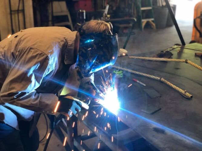 A person wearing protective gear welds metal on a workbench, with sparks flying, in a workshop setting.