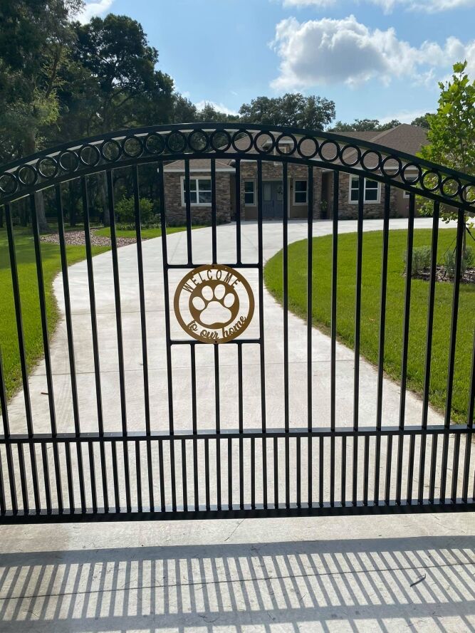 Black metal gate with a gold paw print emblem and Our home, is our home text, in front of a curved driveway leading to a brick house with green lawn and trees.