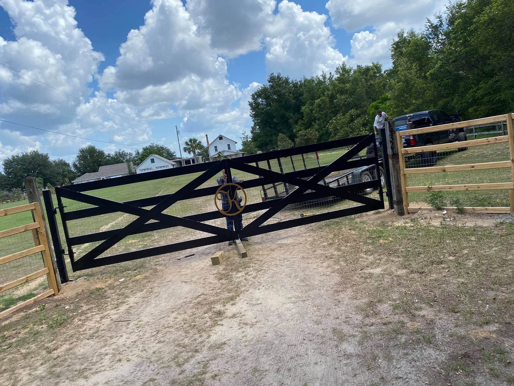 Black double gate with a circular emblem in the center, installed at the entrance of a fenced rural property with a house and trees visible in the background.