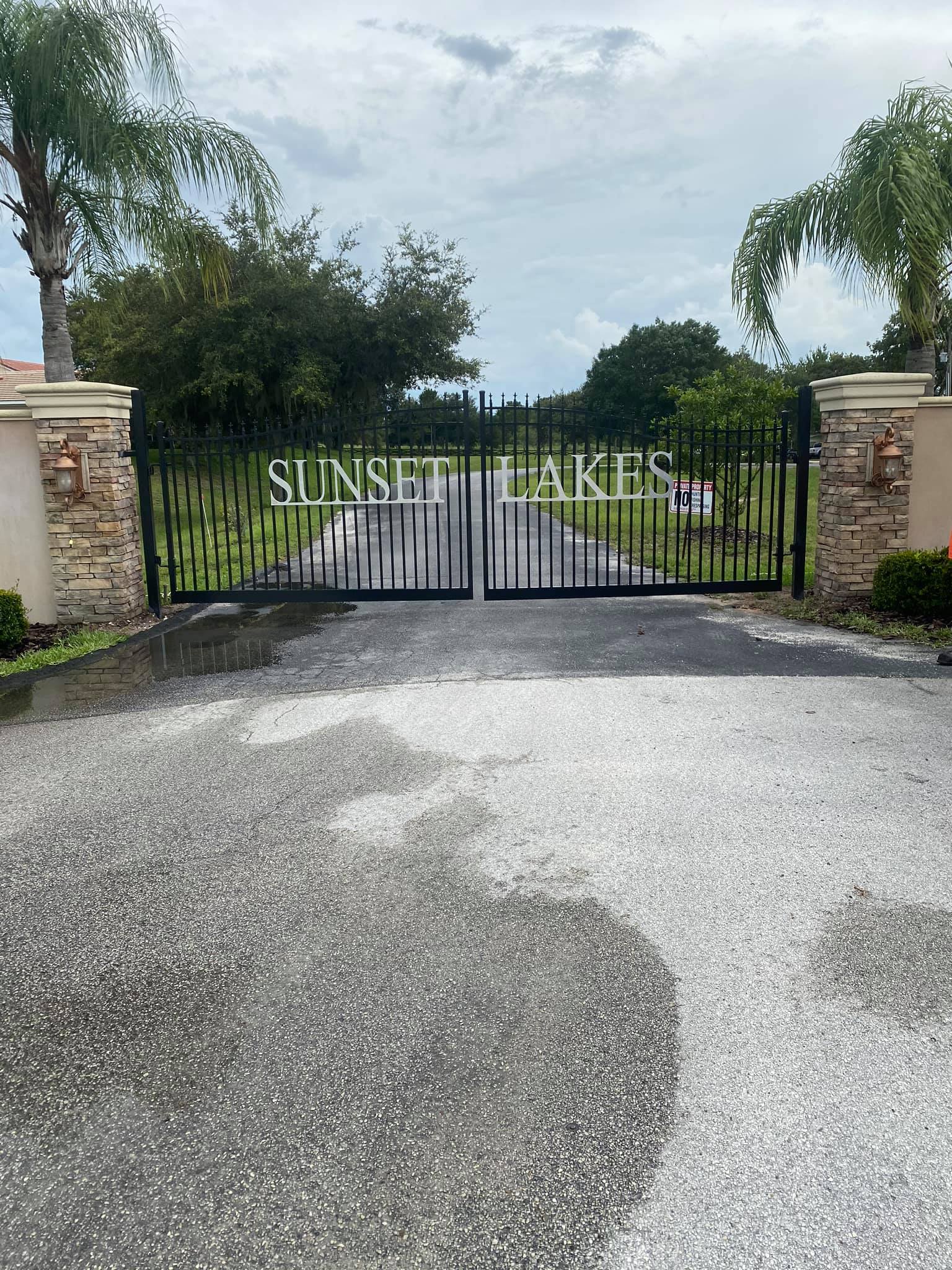 Black metal gates with “SUNSET LAKES” written across, flanked by stone pillars and palm trees, block the entrance to a residential area on a cloudy day.