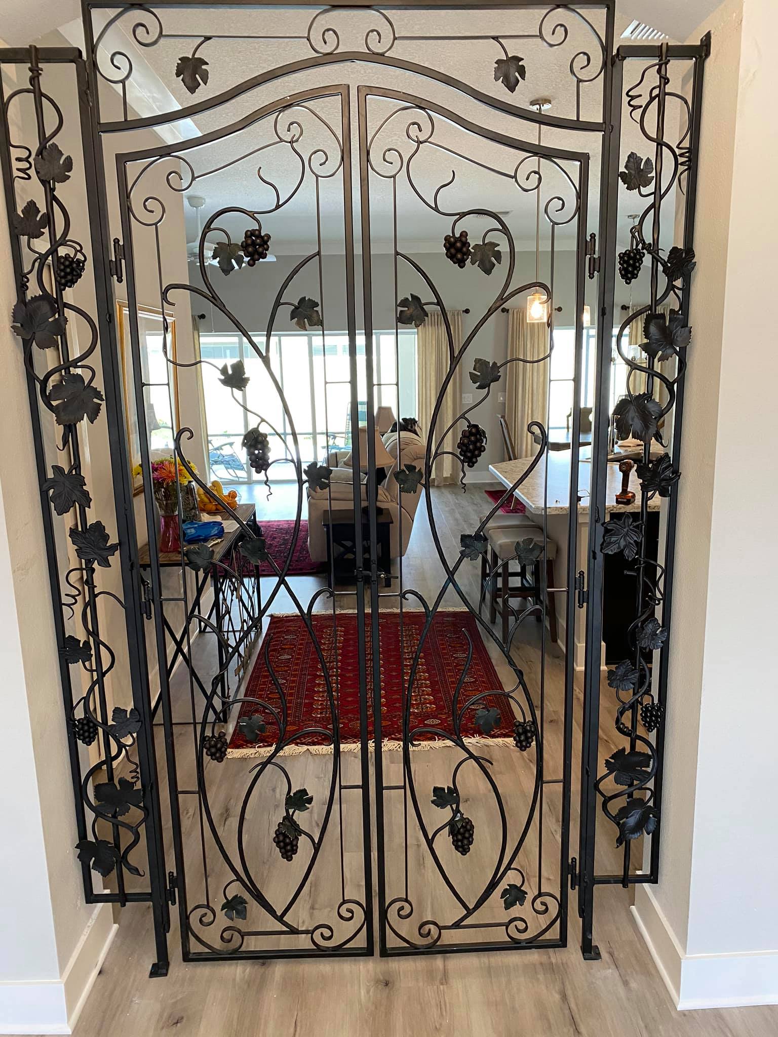 Ornate black metal gate with grapevine and leaf details frames the entrance to a living area with a red rug and light-colored floors.