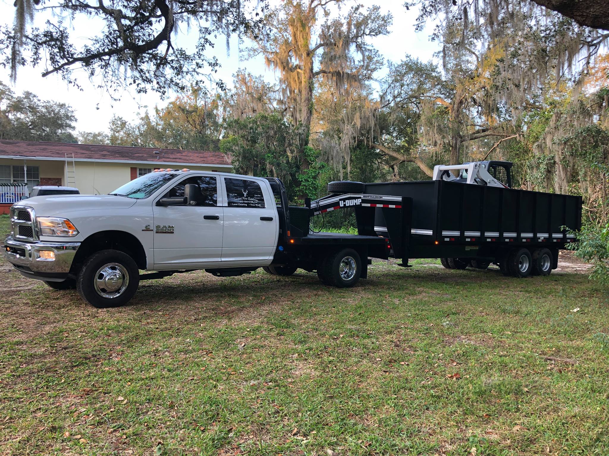A white pickup truck towing a black gooseneck trailer with a skid steer loader parked inside on a grassy yard near a house with trees in the background.