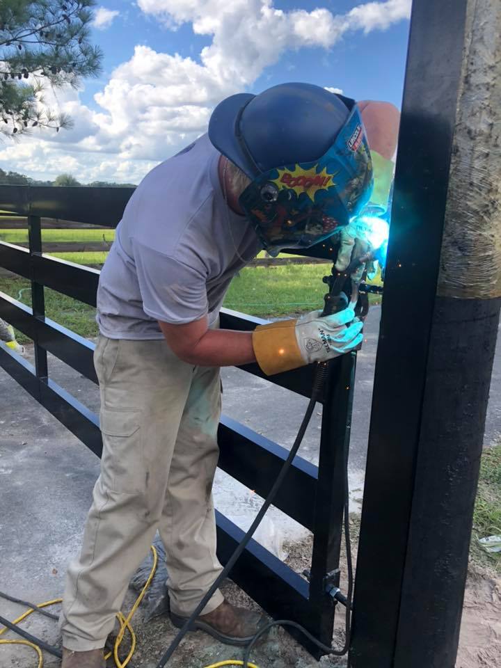 A person wearing protective gear welds a metal gate outdoors on a sunny day.